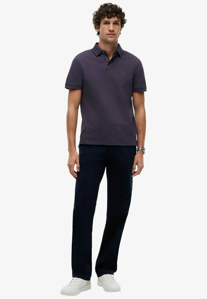 Homme debout portant un polo violet foncé, un pantalon noir, des baskets blanches et une montre-bracelet argentée, sur un fond uni.