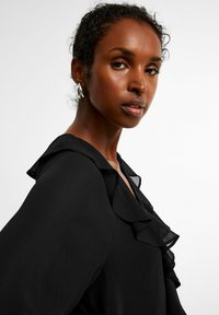 Svart blus med volangkrage, långa ärmar och textil med struktur, där en modell visar upp silverfärgade hoopörhängen.
