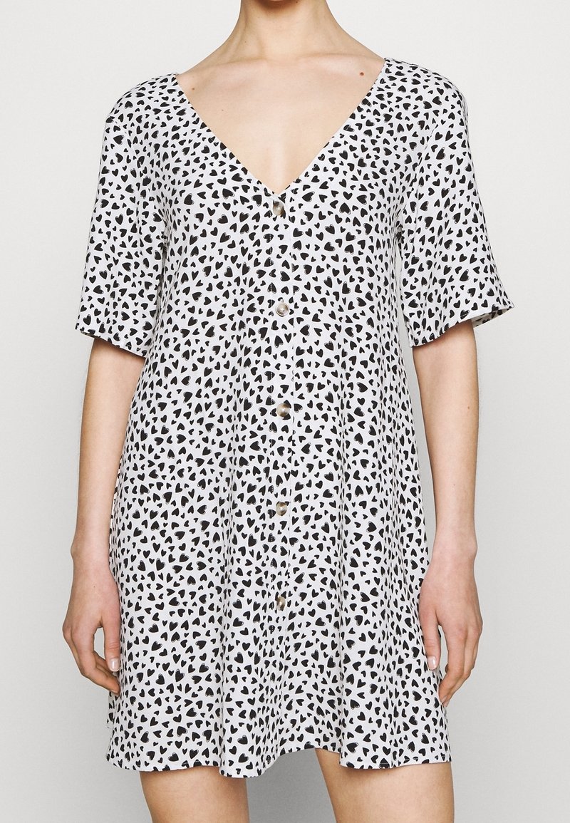 Vestido de manga corta y escote en V en tejido ligero, con un fondo blanco adornado con patrones de corazones negros y detalle de botones en el frente.