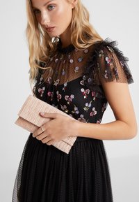 Svart kjole med blomsterbroderi har en gjennomsiktig prikket yoke og volangermarmer. Rosa plisserte clutch bæres i hånden, sølvring på fingeren.