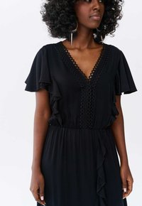 Robe noire avec des manches volantées, encolure en V avec un contour en dentelle circulaire, taille cintrée, détail de volants sur le côté et texture lisse qui tombe.