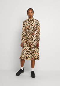 Vestido de padrão de leopardo em bege, com mangas longas, colarinho com botões, cintura cingida e comprimento até à metade da panturrilha. Combinado com ténis pretos.