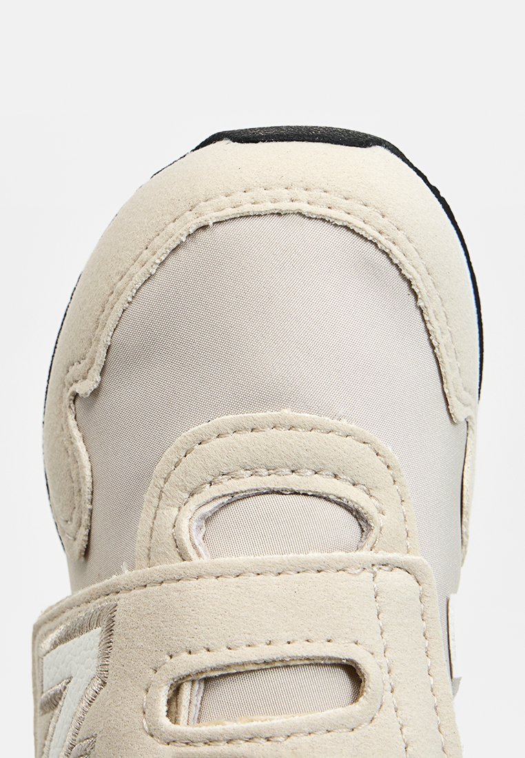 Scarpa sportiva beige con tomaia in tessuto testurizzato, dettagli in suede e suola in gomma nera. Presenta strap in velcro per la chiusura e dettagli di branding.