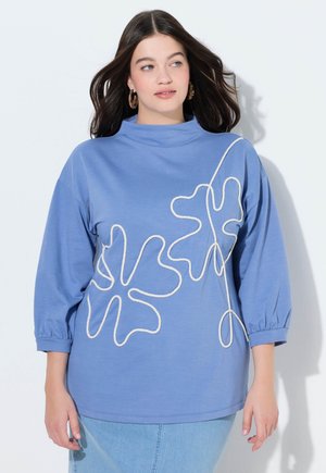 Ung kvinde iført lyseblå langærmet top med hvidt abstrakt blomstermotiv, kombineret med lys denimnederdel, stående mod en hvid væg.