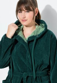 Giacca in corduroy verde con cappuccio foderato in pile di un verde più chiaro e struttura testurizzata. Presenta un cordoncino, una cintura e tasche frontali.