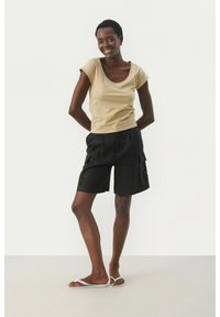 Beige kurzärmliges Oberteil mit Rundhalsausschnitt, kombiniert mit schwarzen Cargo-Shorts mit Seitentaschen. Das Model trägt weiße Flip-Flops.
