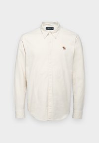 Lys beige skjorte med knaplukning, spidse kraver, lange ærmer og et lille broderet logo af en elg i brune nuancer på venstre bryst. Lavet af bomuldsstof.