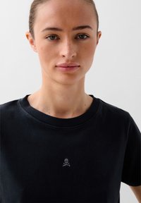 Black cotton crew neck t-shirt featuring a small white embroidered skull and crossbones design on the chest. Smooth texture.
