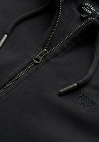 Sudadera con cremallera negra hecha de una tela suave, con un diseño elegante, cremallera metálica con tirador de marca y un sutil detalle de logo.