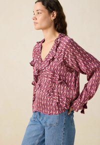 Frau im Profil, die eine lila gemusterte Bluse mit Rüschendetails und blaue Jeans trägt, vor einem einfarbigen beigen Hintergrund.