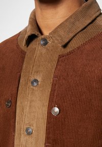 Veste en velours côtelé avec boutons-pression de deux nuances de brun, portée par une personne visible du cou à la poitrine.