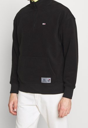Homme portant un sweat-shirt noir à fermeture éclair courte avec poche avant et petit logo, assorti à un pantalon blanc sur fond uni.
