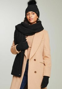 Căciulă tricotată neagră cu pompon, eșarfă din tricot cu model împletit și mănuși asortate. Palton bej, din țesătură texturată. Accesorii casual pentru iarnă.