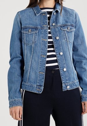 Persoon die een blauwe spijkerjack draagt over een zwart-wit gestreept shirt met een zwarte broek met witte zijstrepen.