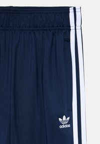 Tamsiai mėlynos sportinės kelnės su tekstūriniu paviršiumi, featuring baltas šonines juosteles ir mažą baltą Adidas logotipą šalia juosmens.