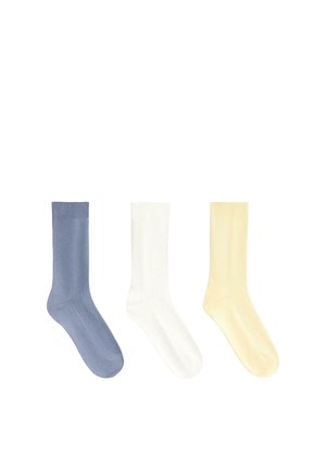 Három egyszerű kötött zokni, kék, fehér és halvány sárga színben, egymás mellett elrendezve, fehér háttéren.