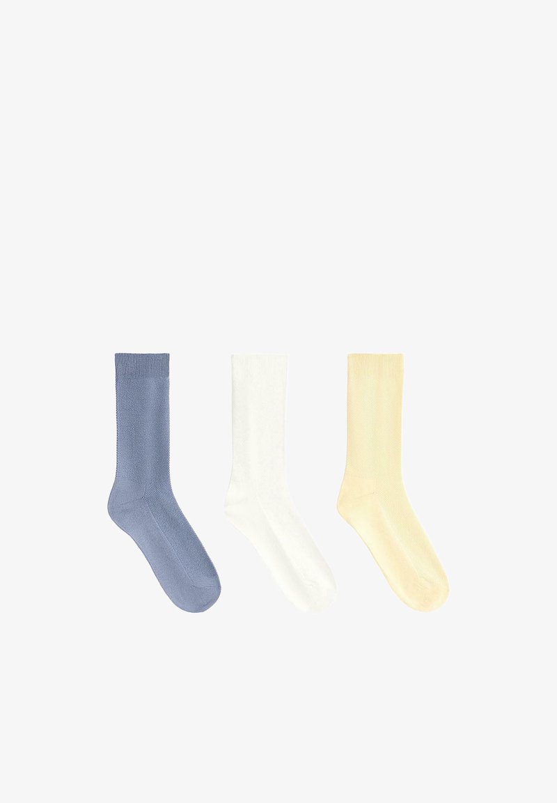 Trois chaussettes unies en maille à côtes, de couleur bleue, blanche et jaune pâle, disposées côte à côte sur un fond blanc.