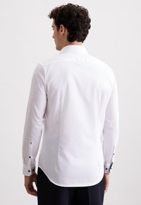 Camicia bianca a maniche lunghe con texture liscia, polsini abbottonati con bottoni neri e orlo curvo. Progettata per una silhouette aderente.