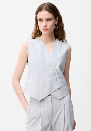 Kvinde med bølget hår iført lysegrå, tyndstribet ærmeløs vest med fire knapper og matchende højttaljede bukser, hænderne i lommerne.