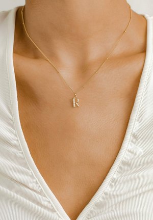 Złoty naszyjnik z teksturowanym zawieszką w kształcie litery "R", z małymi koralikami na łańcuszku, noszony na jasnej bluzce.