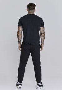 Schwarzes T-Shirt mit kurzen Ärmeln, kombiniert mit schwarzen Sporthosen. Enthält sichtbare Tattoos an den Armen, Schuhe mit weißen Akzenten und schwarzen Sohlen.