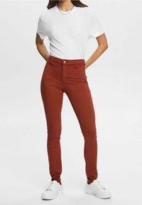 Nainen, jolla on päällään korkeat ruosteenväriset kapeat farkut, valkoinen t-paita, hopeinen ketjukaulakoru ja valkoiset tennarit, poseeraa kädet selän takana.