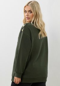 Felpa oversize verde scuro con maniche lunghe, scollo rotondo e dettagli in bottone d'oro sulla spalla. Tessuto morbido.