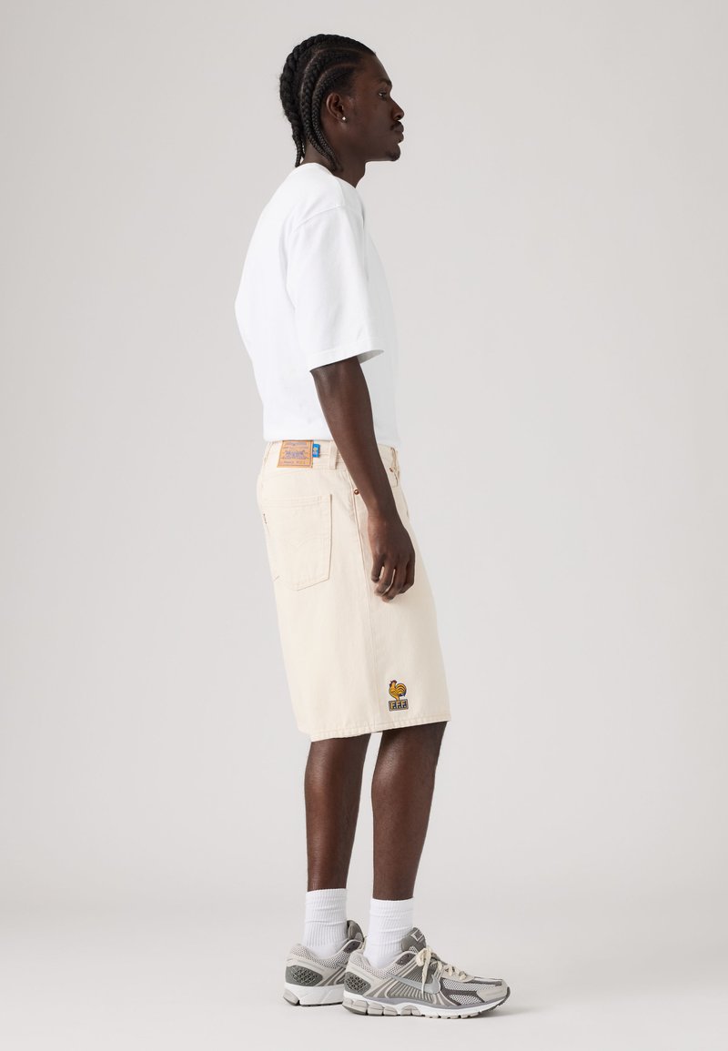 Uomo che indossa una maglietta bianca oversize, pantaloncini crema fino al ginocchio con un logo di gallo, calzini bianchi e scarpe da ginnastica grigie, in piedi di lato.