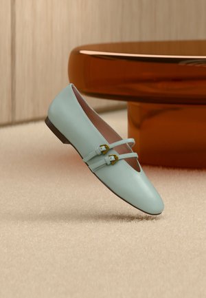 Lichtblauwe Mary Jane platte schoen met dubbele bandjes en gouden gespen op een beige tapijt bij een glanzende bruine ronde tafel.