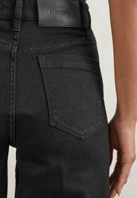 Acercamiento de una persona que lleva puestos unos pantalones de mezclilla negros, mostrando el bolsillo trasero y una etiqueta de cuero sobre la cinturilla.