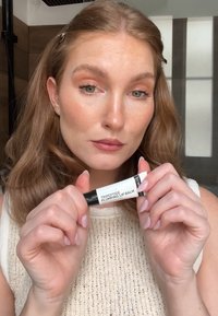 Un tube noir et blanc étiqueté "Baume à lèvres repulpant à tri-peptides." Des mains tiennent le produit devant un fond de couleur neutre.