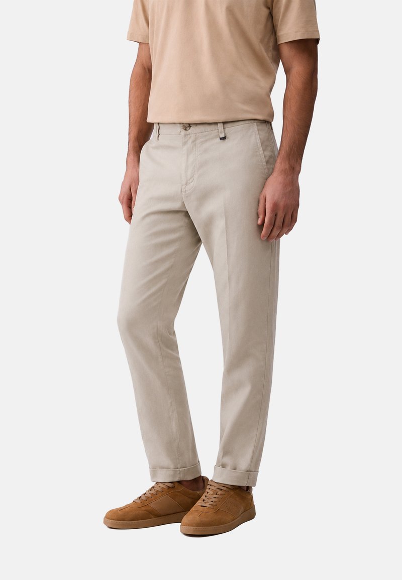 Homme portant un pantalon beige clair retroussé, des baskets beige clair et une chemise beige à manches courtes, debout devant un fond uni.