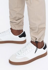 Bílé tenisky s semišovou špičkou, černým akcentem na patě a hnědou gumovou podešví. Texturované panely a tkaničky dotváří casual design.