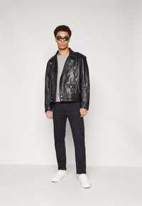 Veste de motard en cuir noir avec des zips argentés, t-shirt gris en dessous, pantalon noir et baskets blanches, portée par une personne debout.