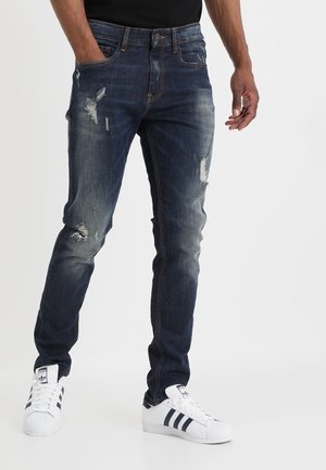 Homme portant un jean skinny bleu foncé usé avec décoloration et des baskets Adidas blanches à rayures noires, debout avec une main dans la poche.