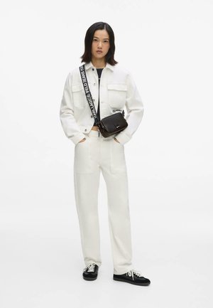 Donna con giacca e pantaloni bianchi, borsa a tracolla nera e sneakers nere, in piedi con le mani in tasca su sfondo bianco.