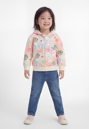 Jeune fille souriante portant un sweat à capuche zippé GAP à motif floral, un jean bleu et des chaussures rose clair, debout sur un fond blanc uni.