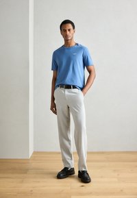 Blauwe katoenen T-shirt met korte mouwen, lichtgrijze op maat gemaakte broek, zwarte riem en chunky zwarte schoenen, staand op een houten vloer tegen een lichte muur.