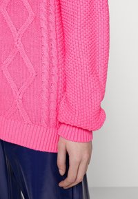 Ljusrosa stickad tröja med texturerade kabelmönster, ribbad nederkant och långa ärmar, tillsammans med glänsande marinblå byxor.