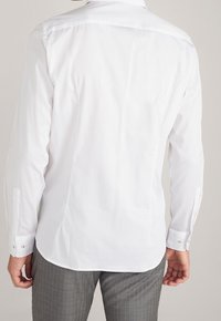 Camisa blanca de manga larga con cuello estándar, tejido de algodón suave y puños abotonados. La vista trasera muestra un diseño sencillo y costuras discretas.