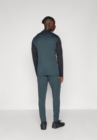 Pánsky športový outfit v tmavej tyrkysovej farbe s čiernym topom s dlhým rukávom. Obsahuje úzke nohavice, rebrované manžety a reflexné detaily.
