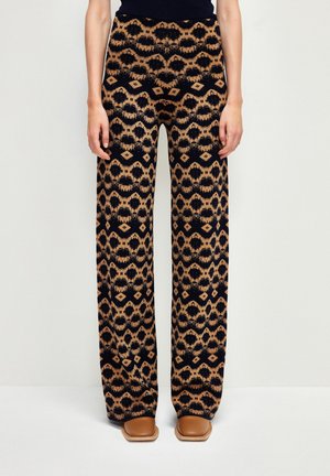 Vrouw die een broek met hoge taille en wijde pijpen draagt, met een zwart-bruine geometrische tribale print en tan kleurige schoenen met een vierkante neus.
