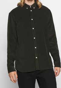 Mand iført en sort kordfløjls skjorte med hvide knapper og sorte bukser, stående foran en ensfarvet baggrund.