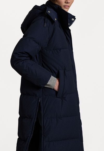 Geacă puffer bleumarin cu glugă, dotată cu fermoar frontal, buzunare laterale și manșete stil rib. Design matlasat cu o siluetă lungă.