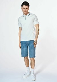 Biała koszulka polo z kontrastowym kołnierzykiem, niebieskie szorty dżinsowe, białe trampki i skarpetki do kostek. Model stoi na jednolitym tle.