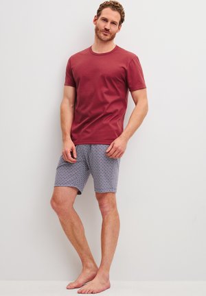 Man staat op blote voeten tegen een witte muur, draagt een rood T-shirt met korte mouwen en blauw korte broek met een geometrisch patroon.