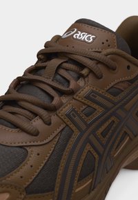 Καφέ αθλητικό παπούτσι ASICS με σκούρο καφέ πλέγμα, λεπτομερείς ραφές και κορδόνια δεμένα πάνω από την επωνυμοδοτούμενη γλωττίδα.