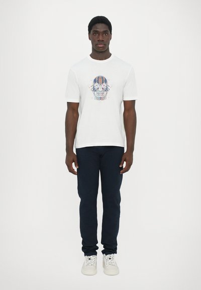 T-shirt en coton blanc avec un graphique de skull coloré, associé à un pantalon bleu marine et des baskets blanches. Design simple avec des manches courtes.