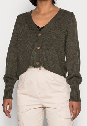 Personne portant un cardigan en maille boutonné couleur olive foncé avec manches longues et un pantalon taille haute beige clair avec poches.