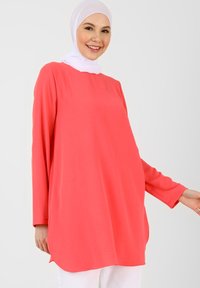 Tunic corallo a maniche lunghe, vestibilità ampia, scollatura rotonda, tessuto morbido con una consistenza liscia. L'orlo ha un lieve scallop, indossata con pantaloni bianchi.